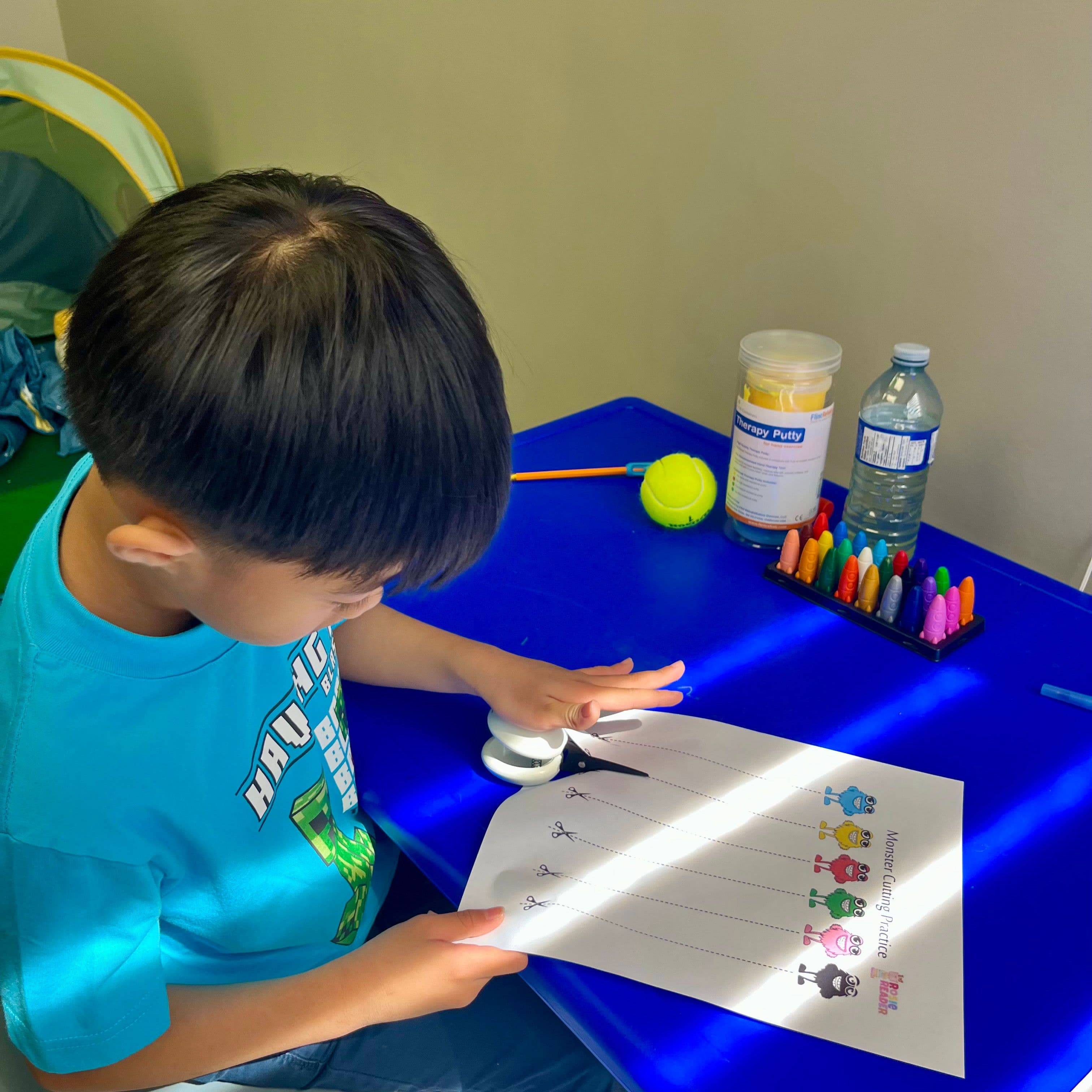 A child at a blue clinic table cutting along dotted lines on a scissor-practice worksheet, with therapy putty and crayons within reach.