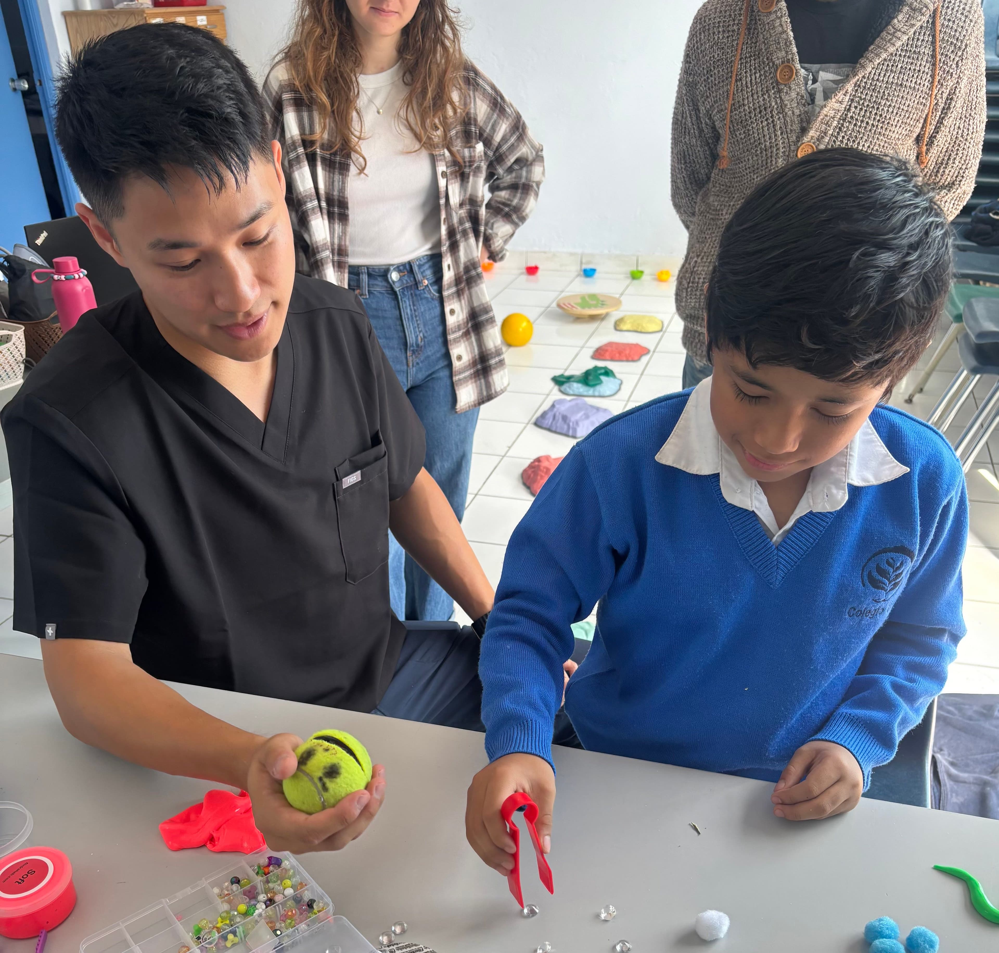 Leo holds up a tennis ball while a boy in school uniform uses red tweezers to pick up beads, with two caregivers watching from behind.