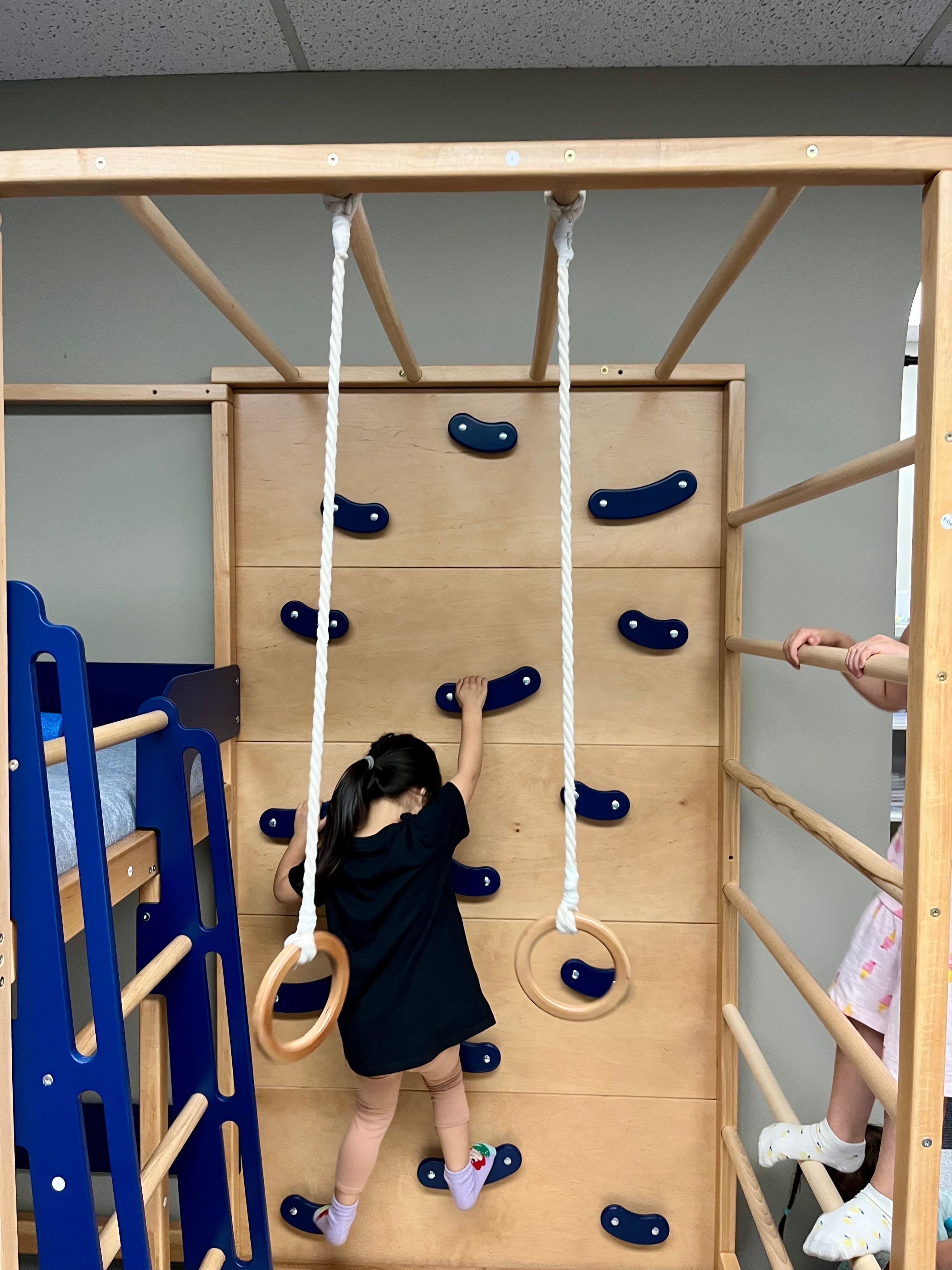 A child reaching for the next handhold on the clinic's wooden climbing wall, seen from behind, with a second child on the rungs alongside.
