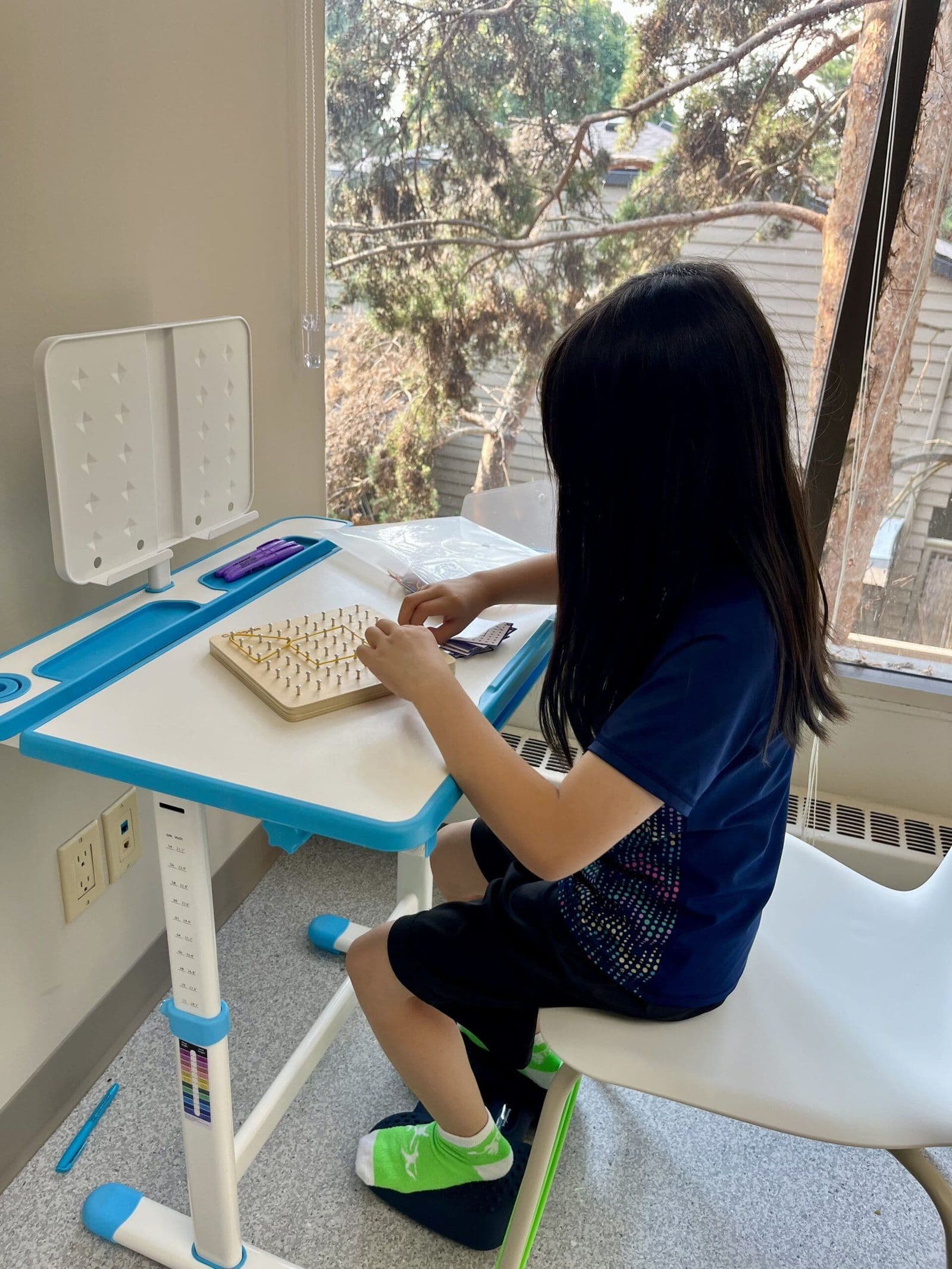 A child uses a pegboard at an adjustable table by a window.