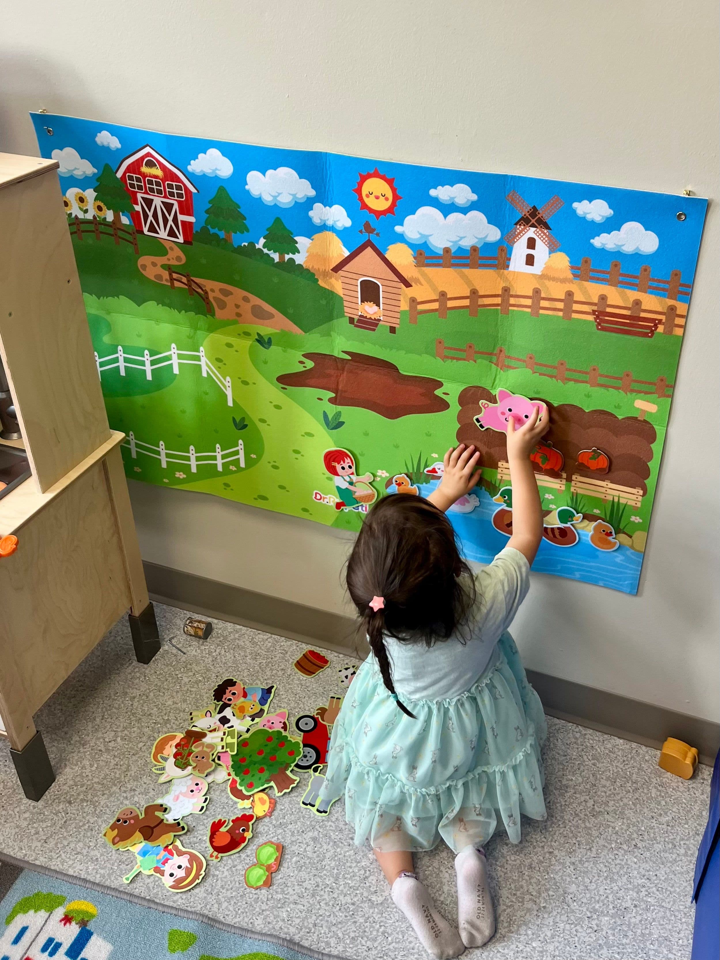 A small child kneeling to press a felt pig onto a farm scene on the wall, with the rest of the felt pieces spread out on the floor.