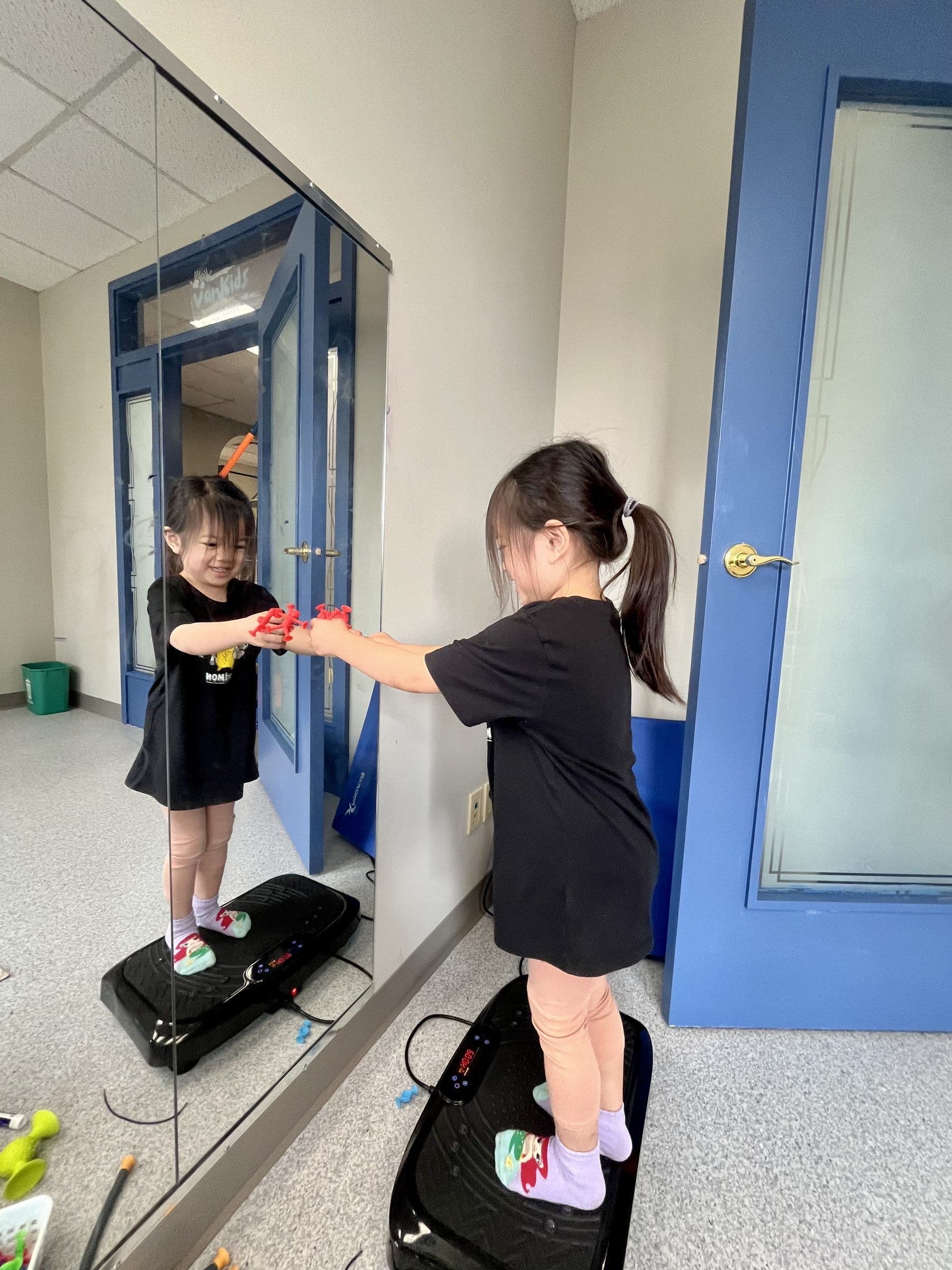 A child stands on a vibration plate at a mirror, stretching a resistance toy between both hands.