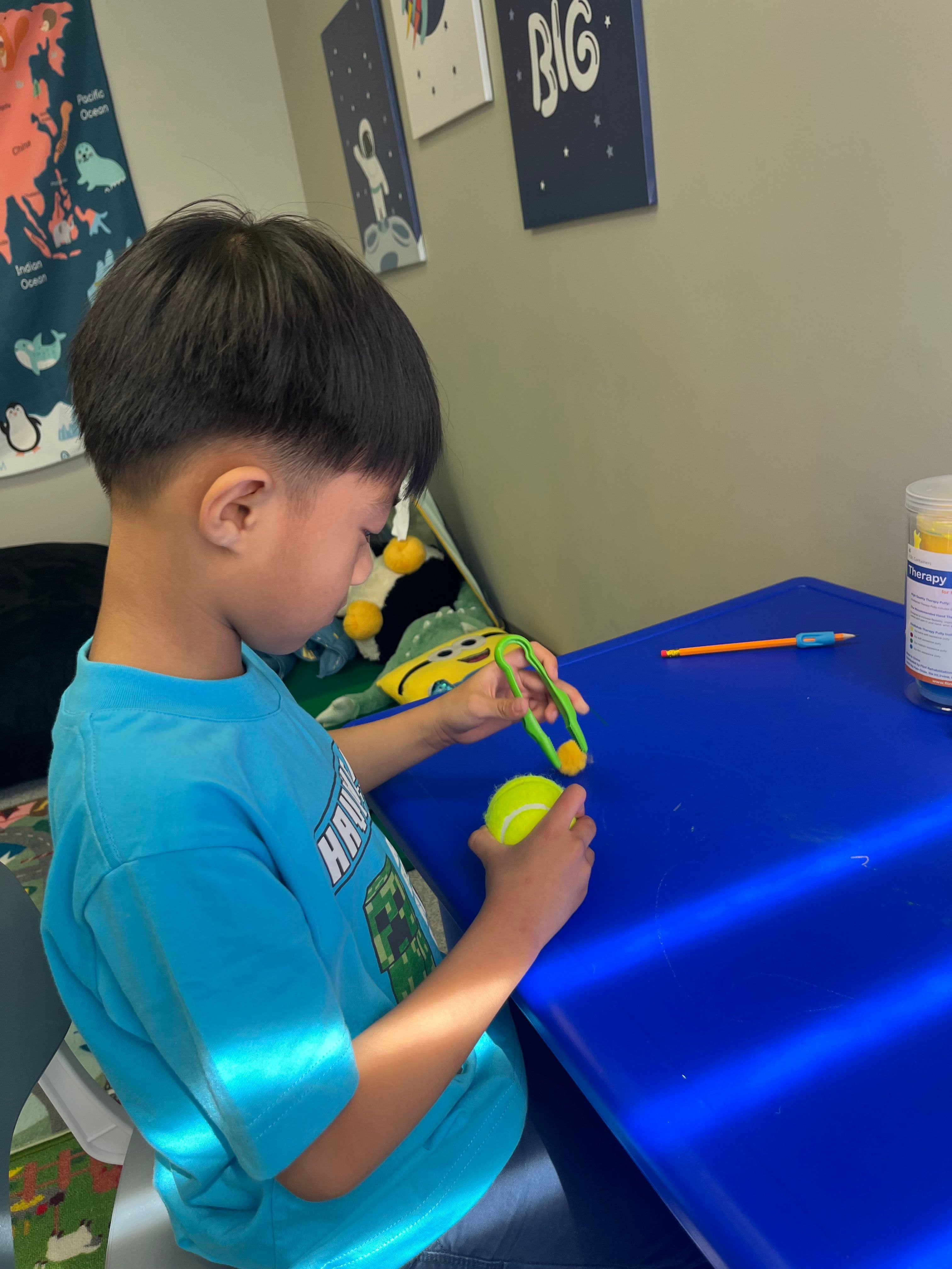 A boy using green tongs to lift a pom-pom while holding a tennis ball in his other hand, working at the blue therapy table.