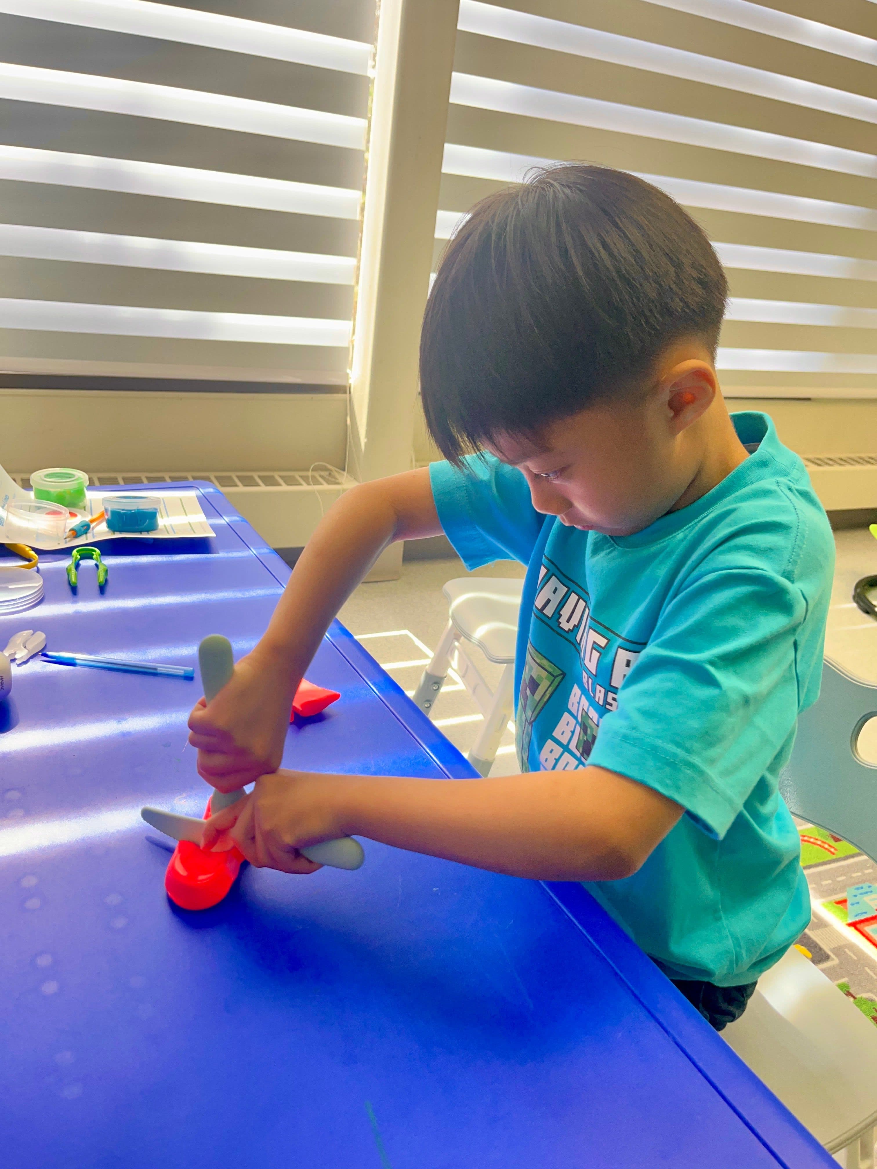 A child gripping scissors two-handed to cut a piece of red therapy putty at a blue therapy table.
