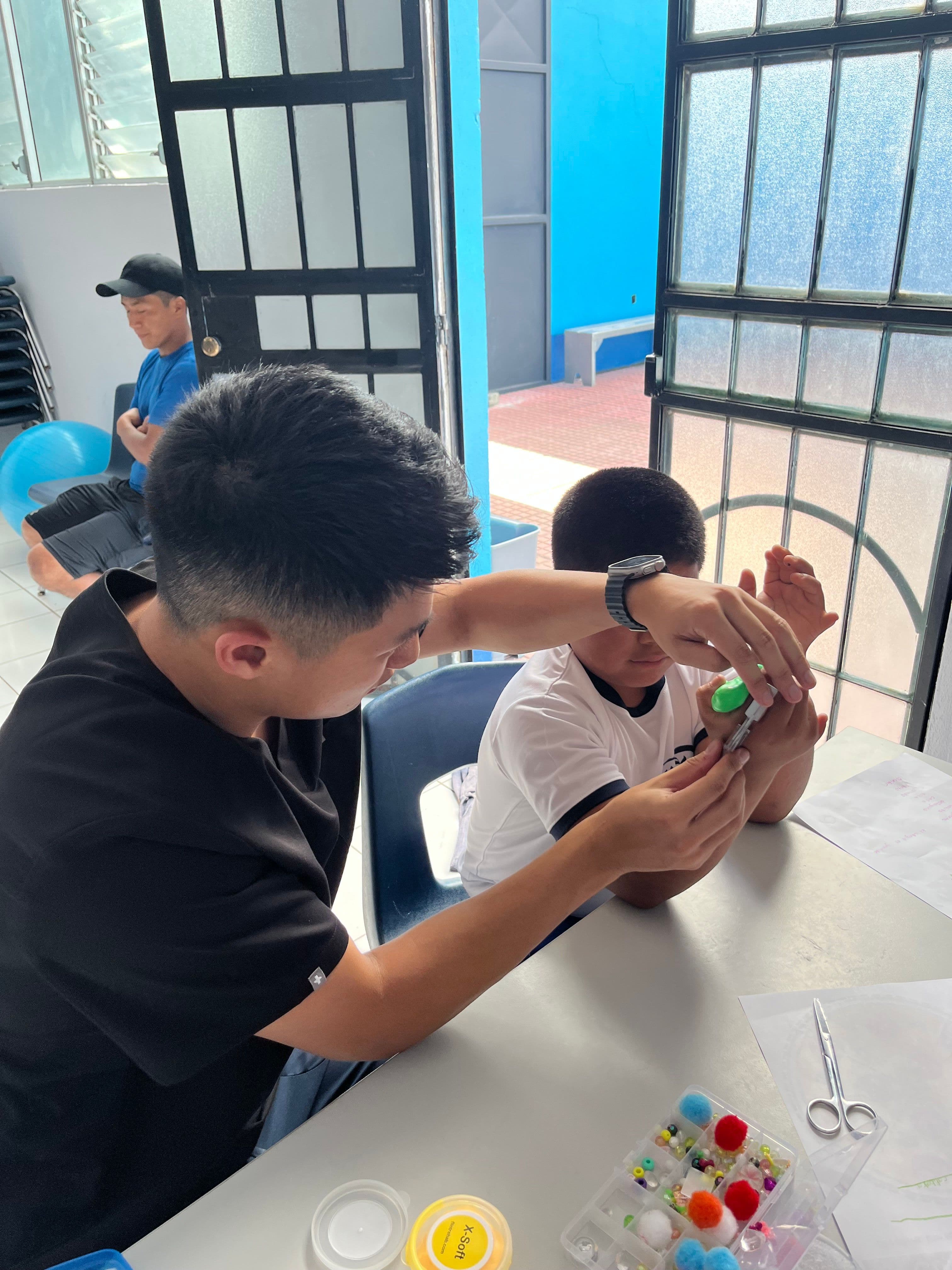 Leo steadies a boy's hands as he pinches a small object with tweezers, at a table laid out with beads and scissors.