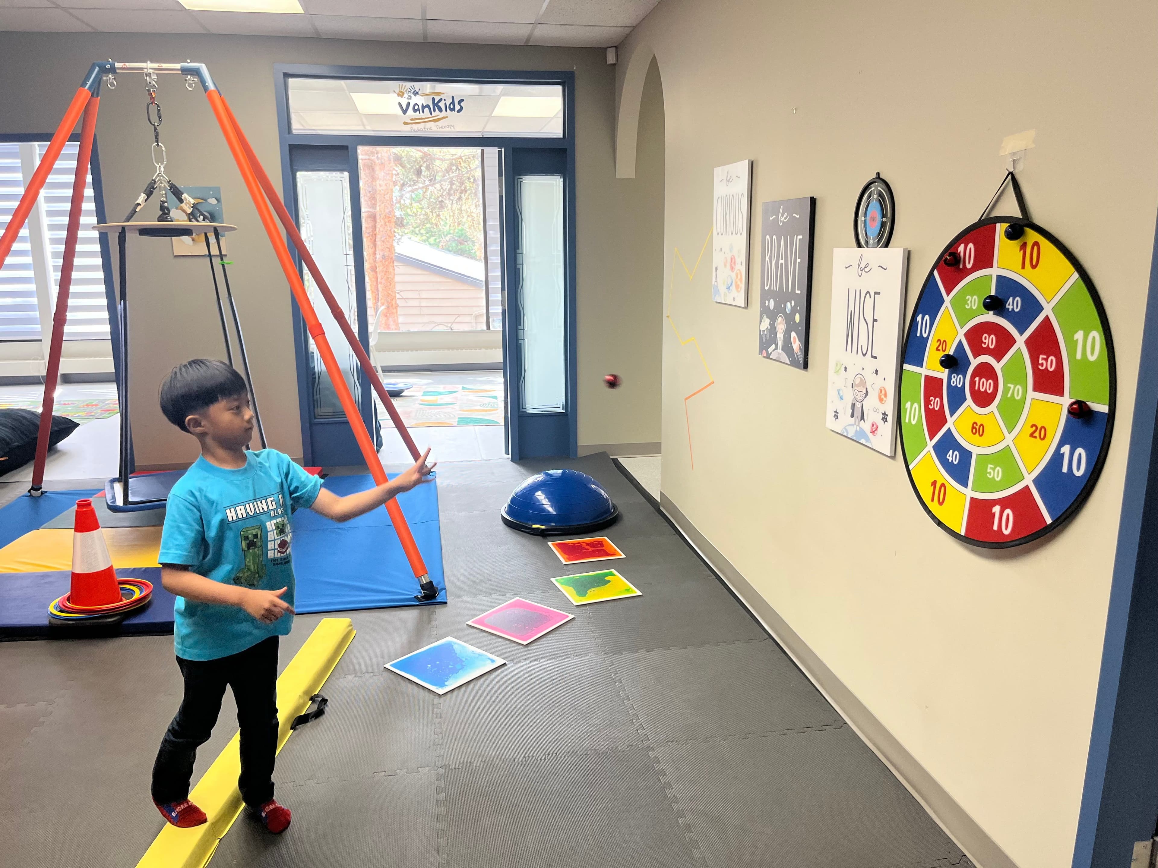 A boy stepping along a yellow balance beam in the gross motor room, with the platform swing, sensory stepping tiles and a target board around him.