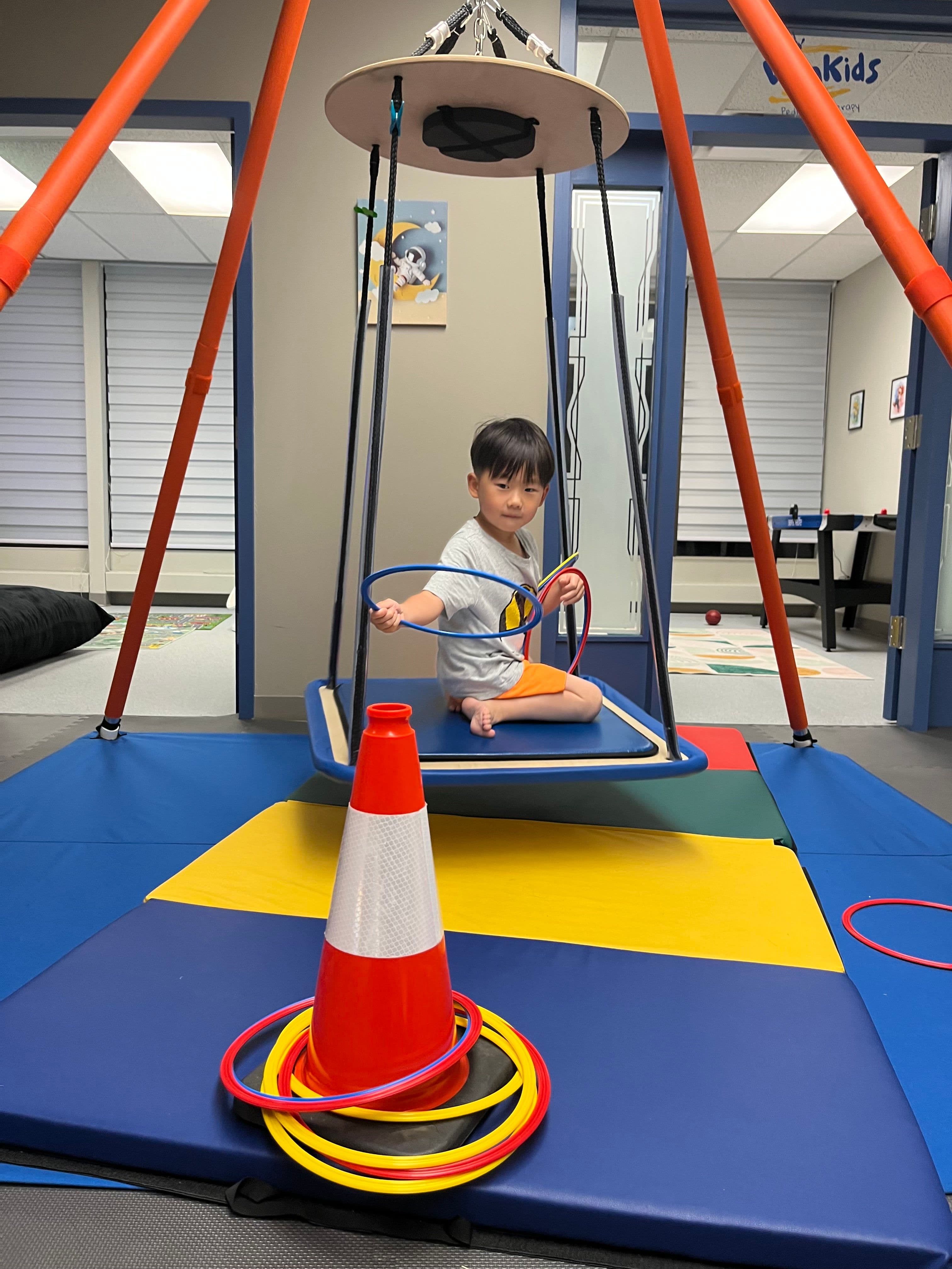 A young boy sitting cross-legged on the platform swing, holding a hoop in each hand, looking towards the camera.
