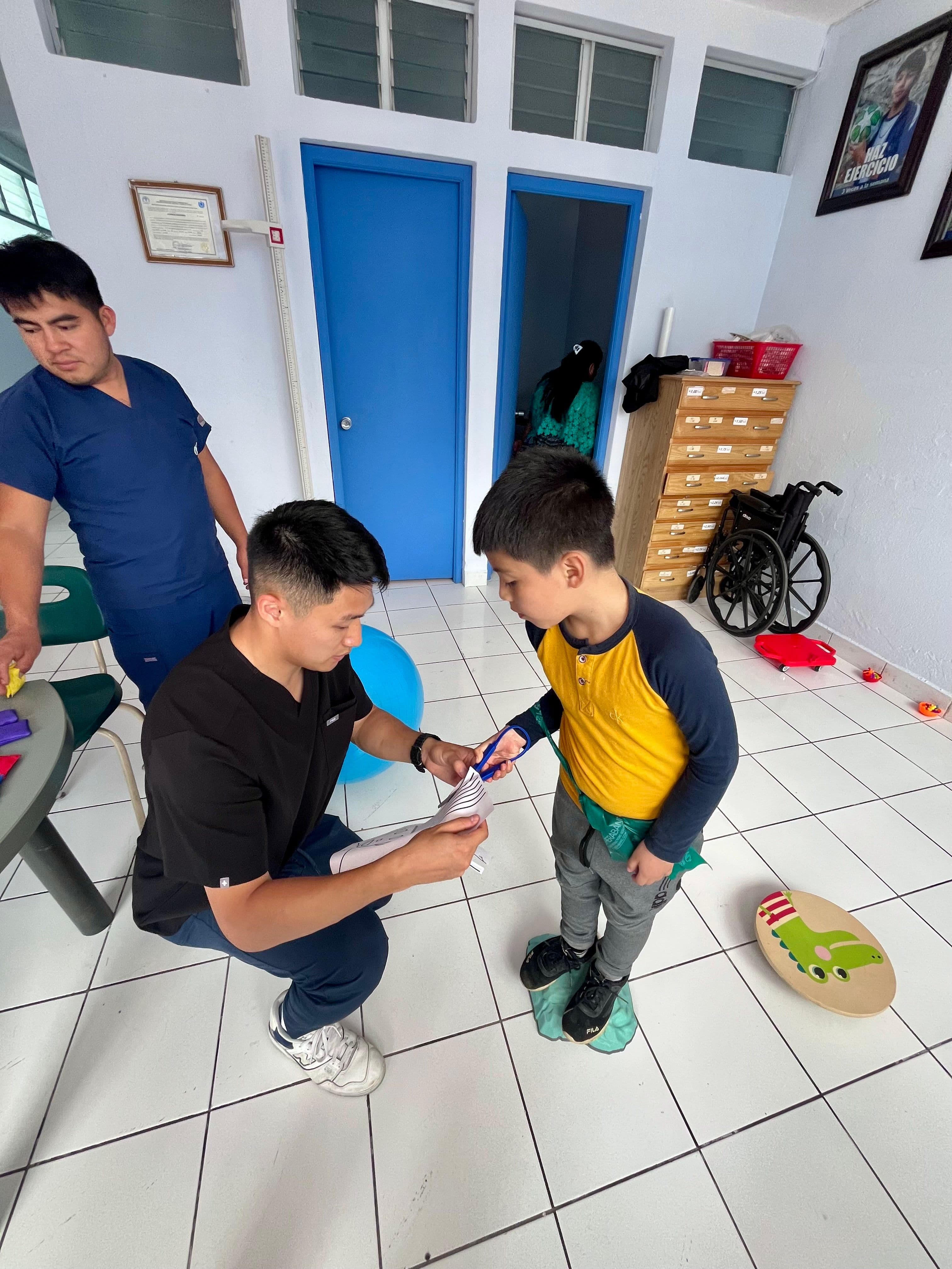 Leo crouches to a boy's eye level to hand him a pair of blue scissors, while a local occupational therapy assistant looks on in the Tactic clinic room.