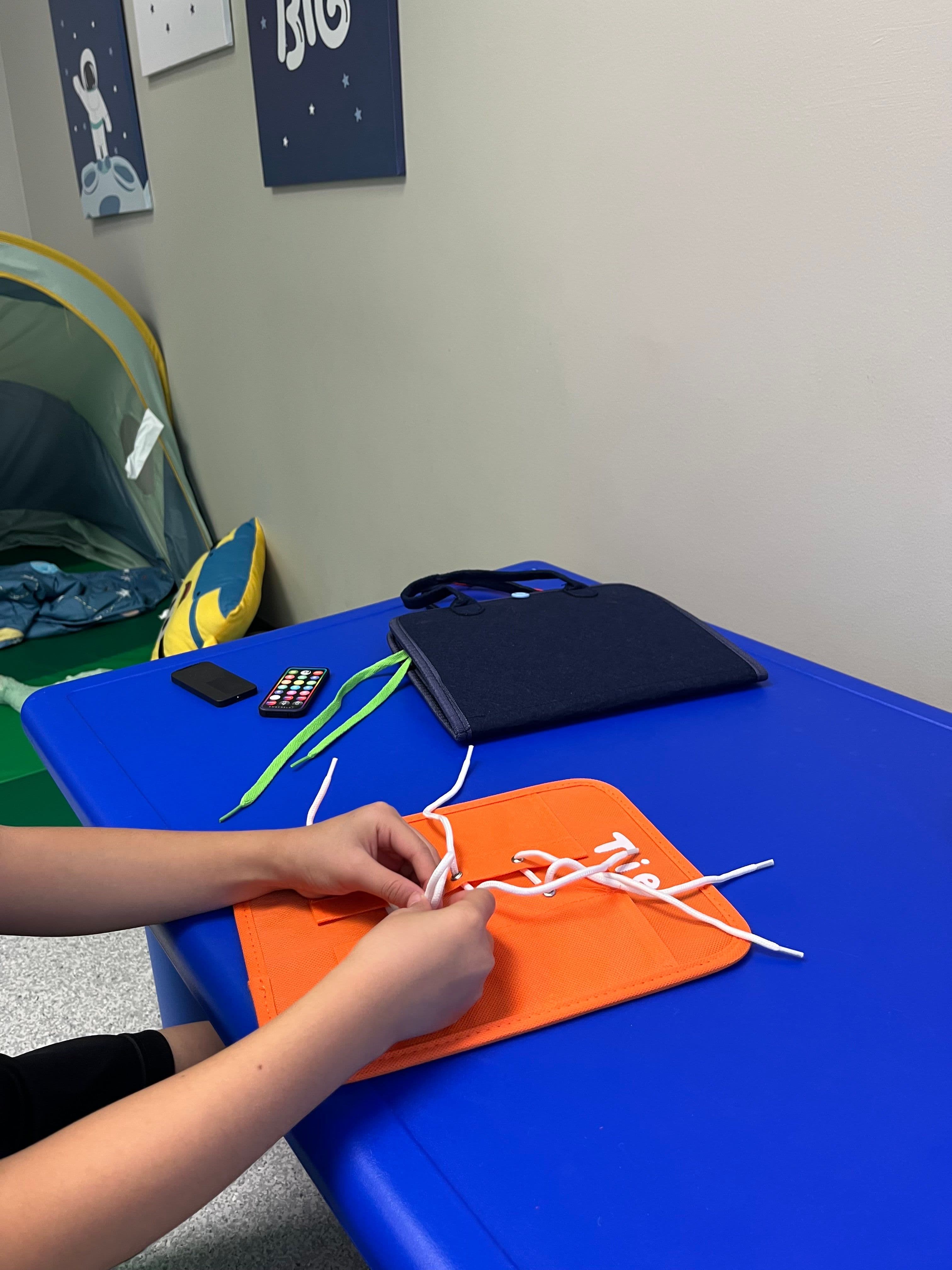 A child's hands threading a white lace through an orange shoe-tying practice board on the blue therapy table.